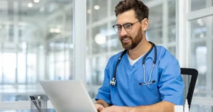 A imagem mostra um veterinário sorridente usando jaleco azul e um estetoscópio no pescoço enquanto trabalha em um laptop. Ele está em um ambiente moderno, com vidro e iluminação natural ao fundo, sugerindo uma clínica ou escritório bem estruturado. O profissional parece concentrado e engajado, transmitindo uma imagem de confiança e profissionalismo, possivelmente relacionado ao marketing veterinário ou gestão de clínica.