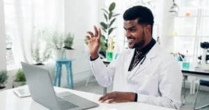 A imagem mostra um veterinário sorridente vestindo um jaleco branco e um estetoscópio ao redor do pescoço, enquanto interage por meio de uma videochamada em um laptop. Ele está sentado em um ambiente bem iluminado, com diversas plantas e equipamentos científicos ao fundo, sugerindo um consultório ou laboratório moderno. Sua expressão amigável e postura engajada destacam a importância da comunicação na área veterinária, possivelmente relacionada ao atendimento remoto ou à troca de informações com colegas e clientes.
